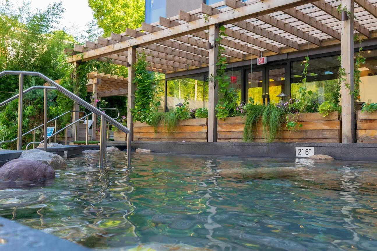 Heated carbon-rich bath at SoJo Spa Club, boulders for sitting, surrounded by lush greenery and flowers. Steps lead to pool entrance. Serene atmosphere.