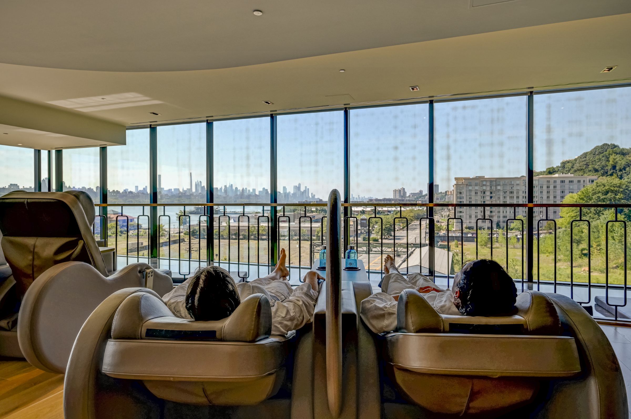 "Two women reclining in comfortable chairs in the relaxation zone of SoJo Spa Club. Both are wearing spa robes and drinking out of SoJo water bottles.