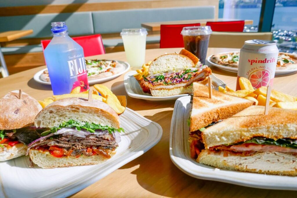 Sandwiches and pizzas sit on a table with a few drinks on a wood table in the SoJo Food Hall